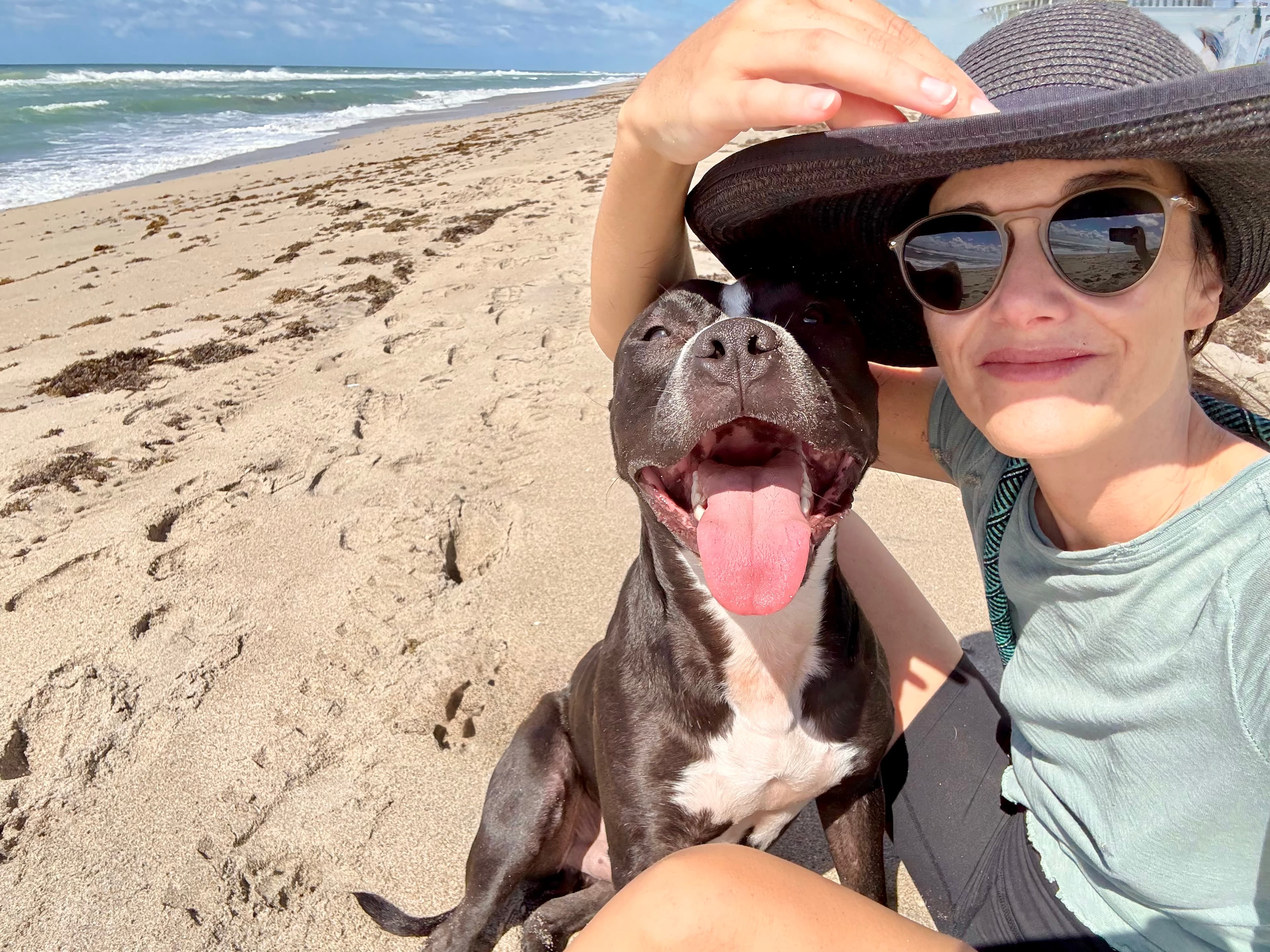 Woman in sun hat takes a selfie with her happy, panting dog on a beach.